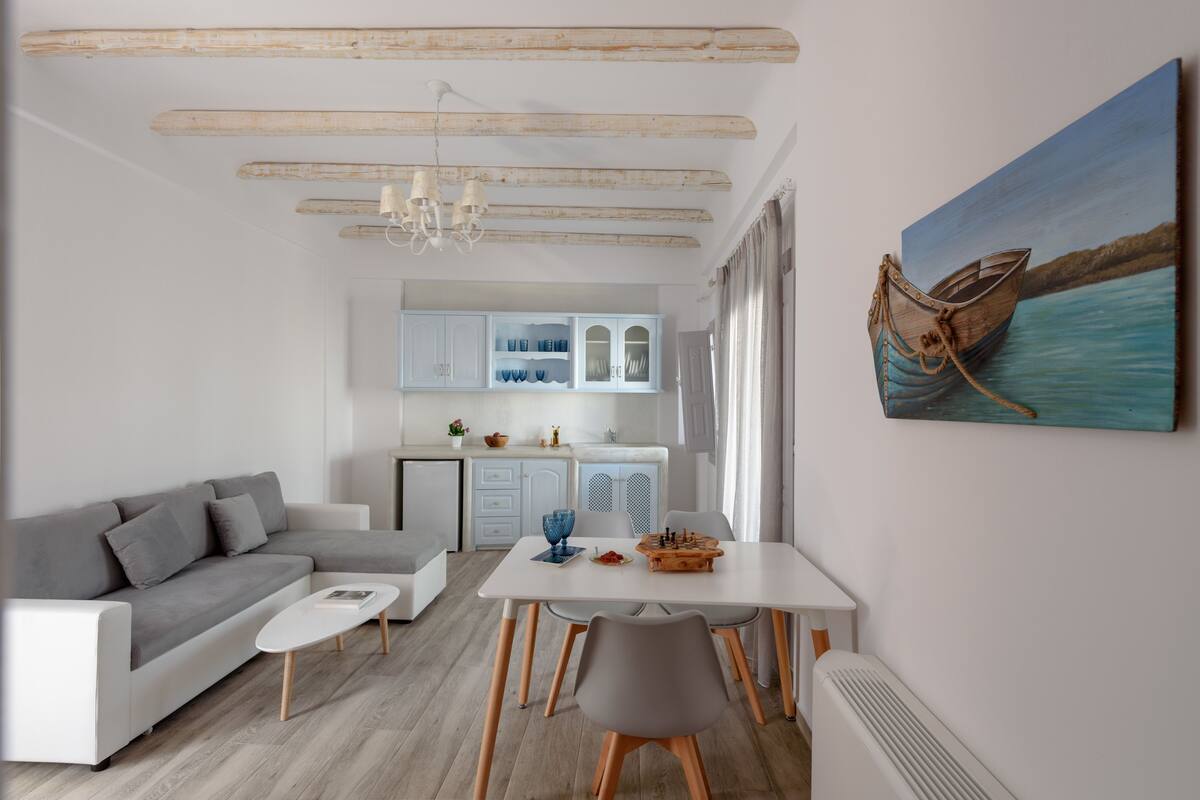 A modern living room with a grey sofa, a white coffee table, and a dining area with a white table and grey chairs. A painting of a boat on water is on the wall.