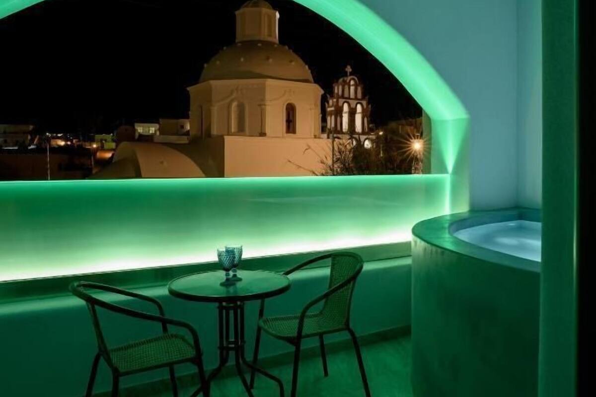 A rooftop terrace with a table and chairs, a hot tub, and a view of a church at night.