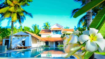 A poolside view of a resort with a clear blue sky, palm trees, and a building with a red roof.
