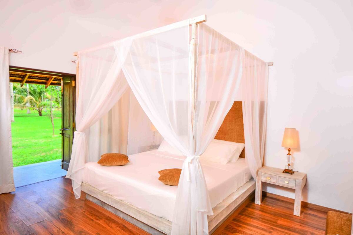 A four-poster bed with white bedding and a canopy, a bedside table with a lamp, and a view of a green outdoor area.
