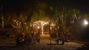 Un espace de restauration situé au bord de la plage, avec des tables et des chaises, entouré de palmiers et illuminé de guirlandes lumineuses.