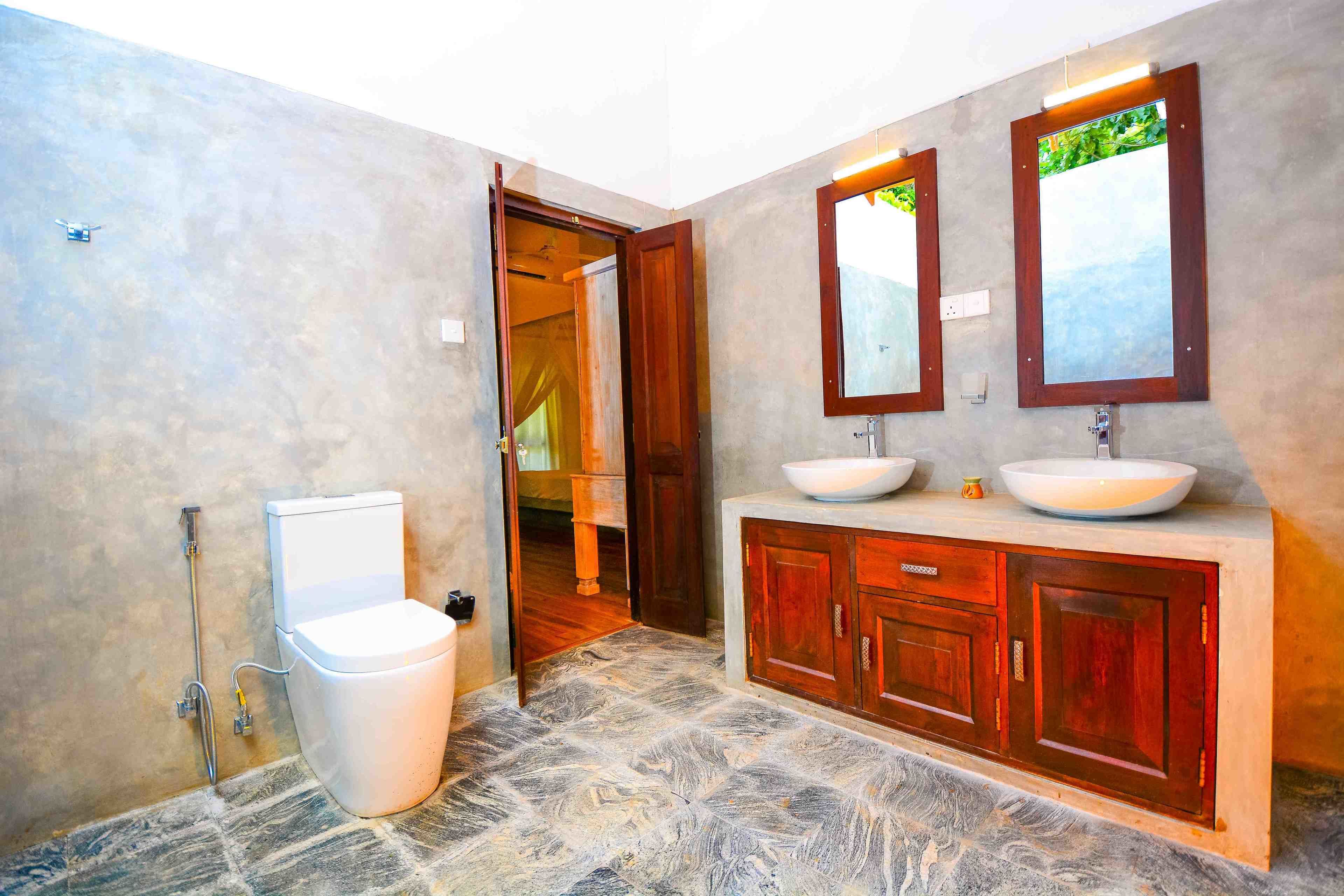 Een badkamer met een stenen vloer, een wit toilet, een houten wastafelmeubel met twee wastafels en een spiegel erboven.