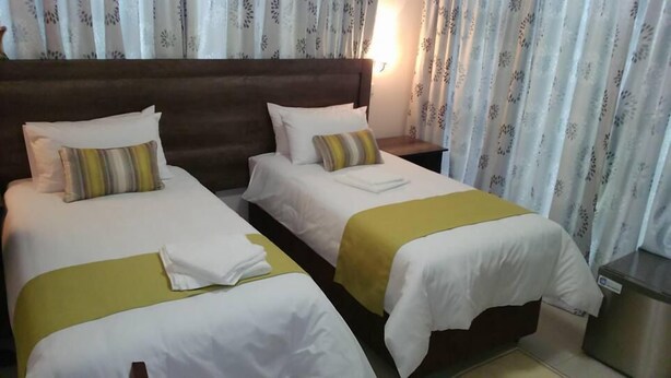 Habitación de hotel con dos camas, cada una con ropa blanca y una manta amarilla. Tiene un cabecero de madera, una mesita de noche con lámpara y una ventana con cortinas.
