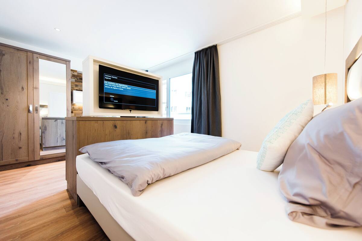 A modern hotel room with a bed, a flat-screen TV mounted on the wall, wooden flooring, and a large window with curtains.