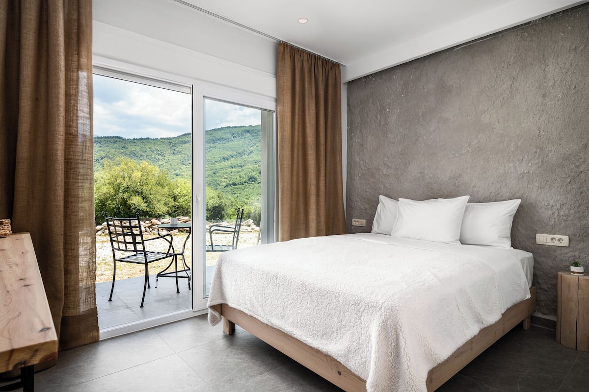 Une chambre avec un grand lit, une vue sur une forêt par une porte coulissante et une petite table avec des chaises.