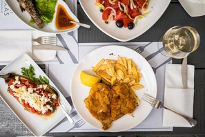 A table with various dishes including fish, fries, and a salad, along with a glass of wine. - Gefiri Plakas Hotel (North Tzoumerka)