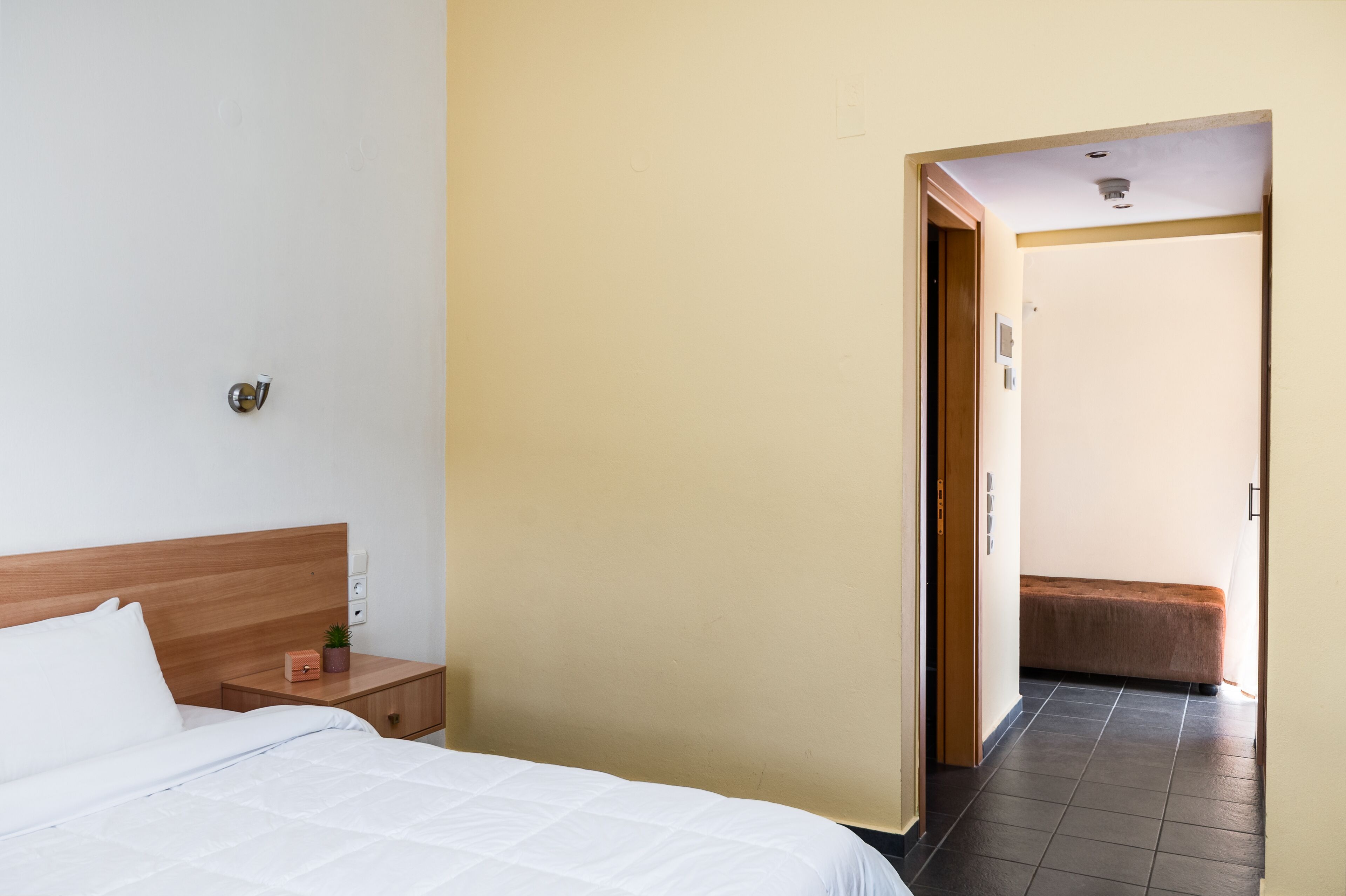 A hotel room with a bed, bedside table, and a bench in the hallway.