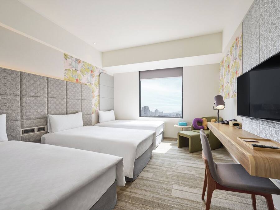 A hotel room with two beds, a desk with a chair, a TV, and a window with a city view.
