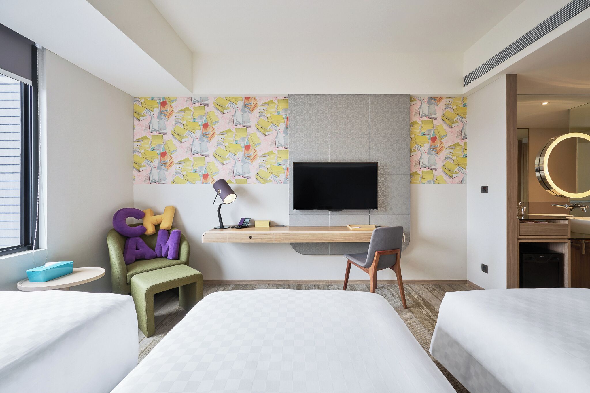 A hotel room with two beds, a desk with a TV, a chair, a small table, and a bathroom with a mirror.
