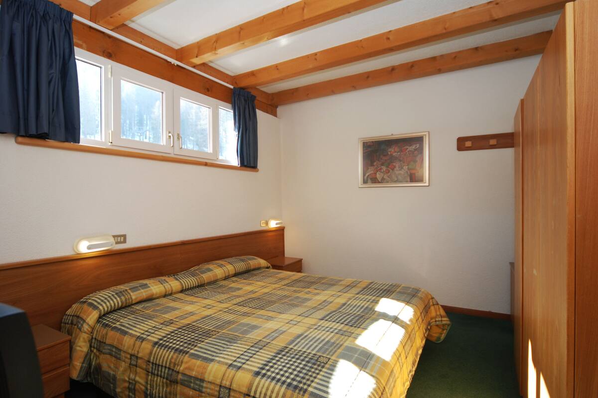 A bedroom with a bed, wooden headboard, a window with curtains, a painting on the wall, and a wooden wardrobe.