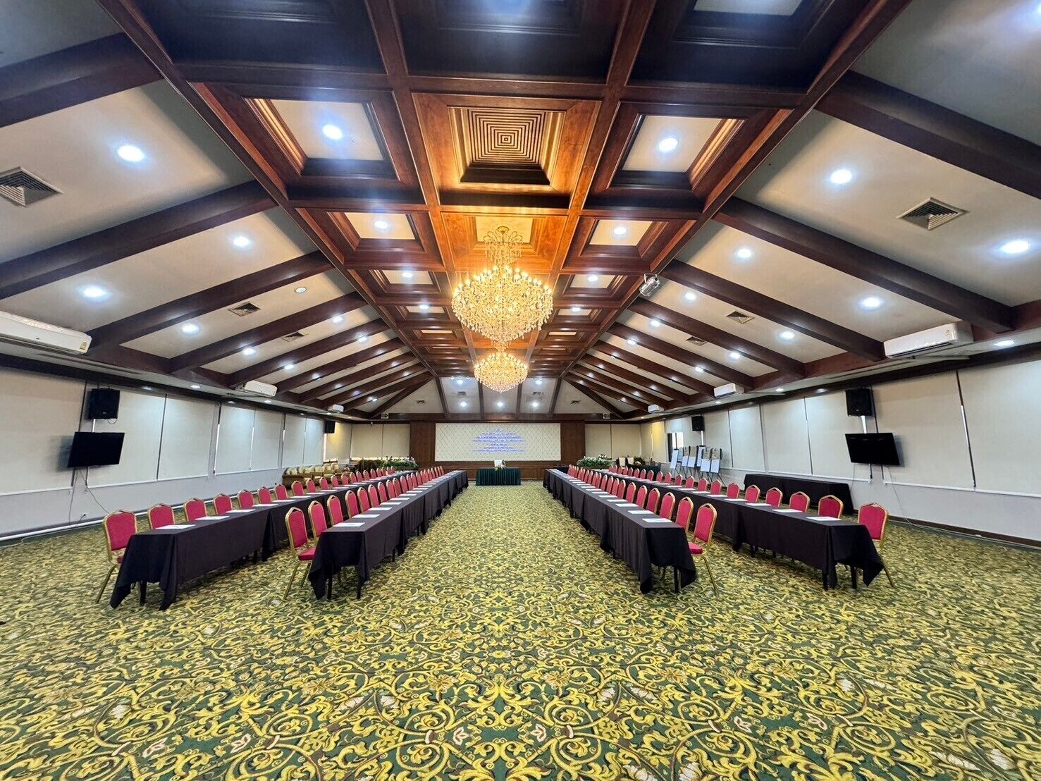 Een congreszaal met een kamerbreed tapijt, lange tafels met stoelen, een kroonluchter en een projectiescherm.
