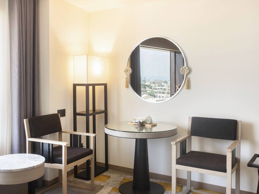 A hotel room with two chairs, a small table, a round mirror, and a view of the city through a window.