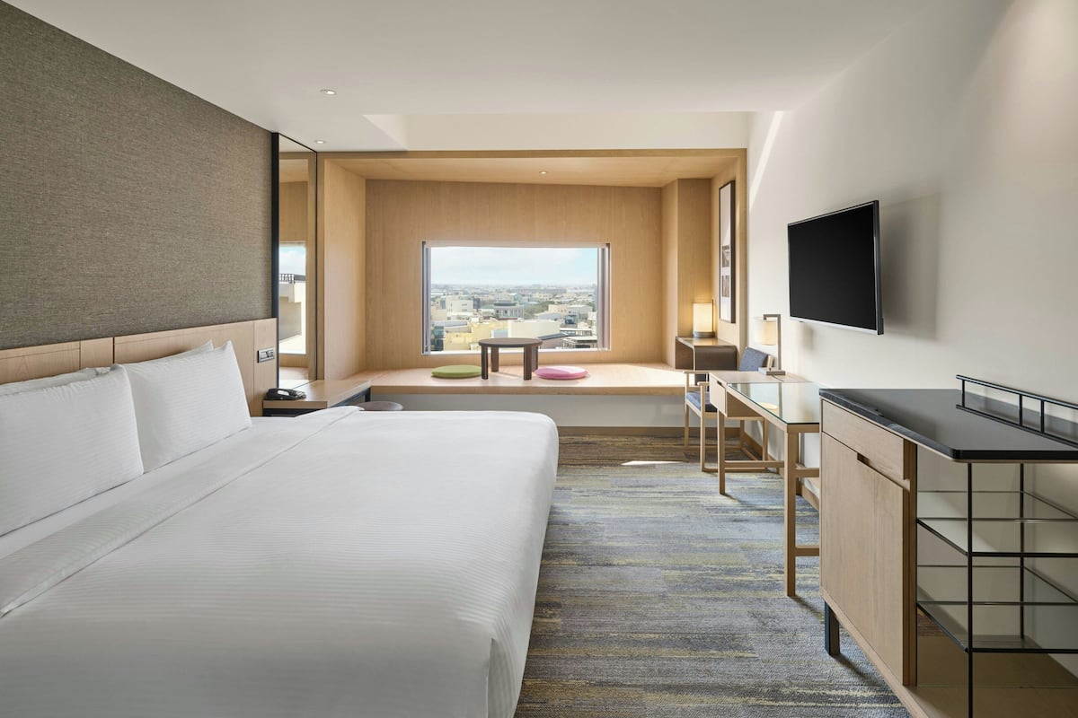 A modern hotel room with a large bed, a desk, a TV, and a view of the city.