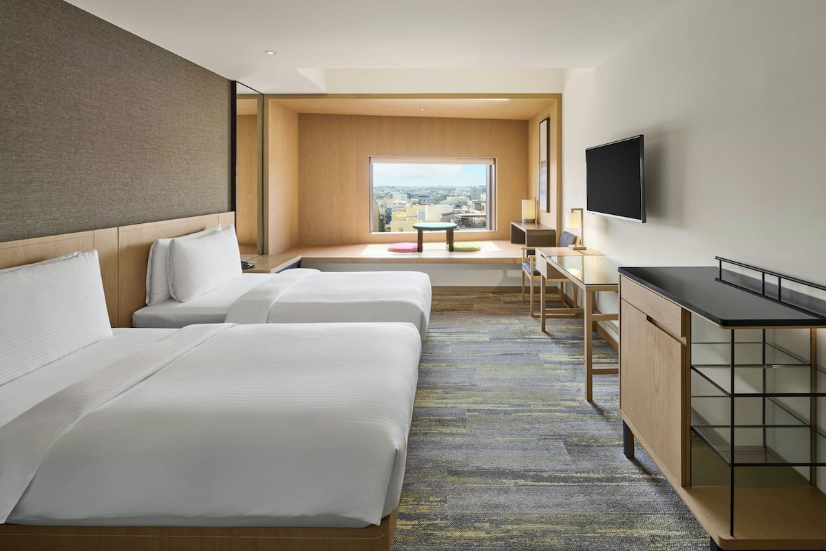 A hotel room with two beds, a desk, a TV, and a large window with a city view.