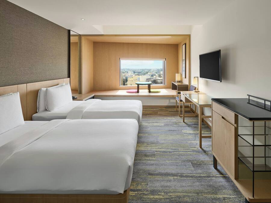 Quarto de hotel com duas camas, uma escrivaninha, televisão e uma grande janela com vista para a cidade.