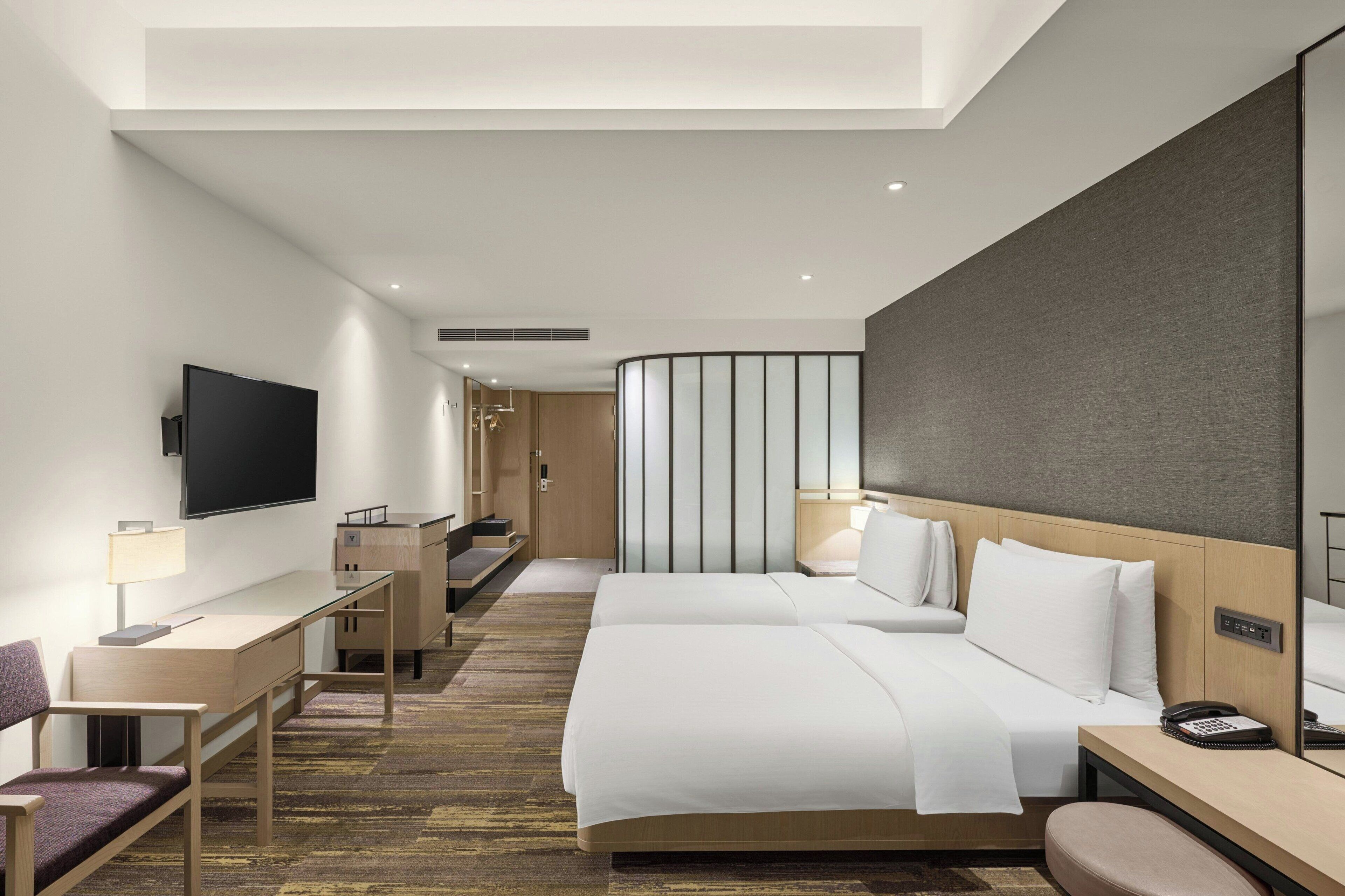 A modern hotel room with a flat-screen TV, a desk with a lamp, two beds, a mirror, and a small bathroom area.