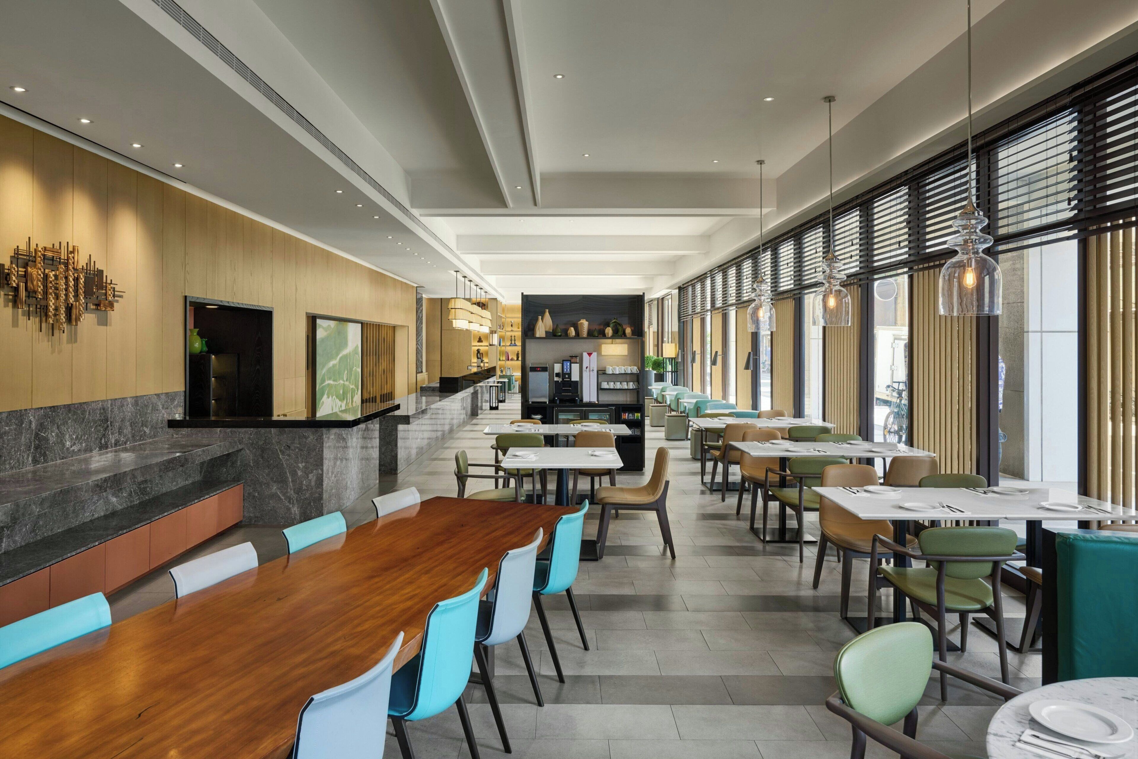 Un restaurante moderno con una larga mesa de madera, sillas azules y verdes, y una zona de cocina al fondo.