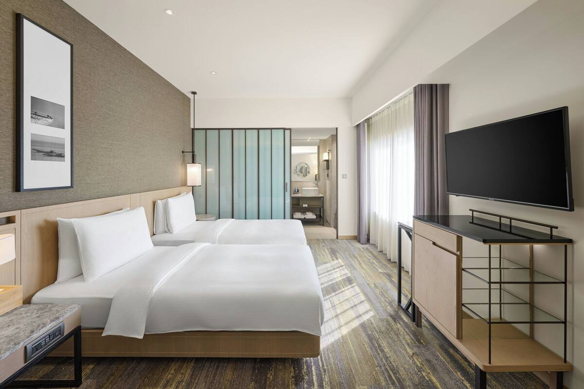 A hotel room with two beds, a TV, a desk, and a bathroom visible through an open door.