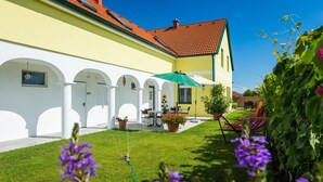 A yellow building with a red-tiled roof, white arches, a patio with a table and chairs, and a green umbrella. - Appartmenthaus Freiler (Morbisch am See)