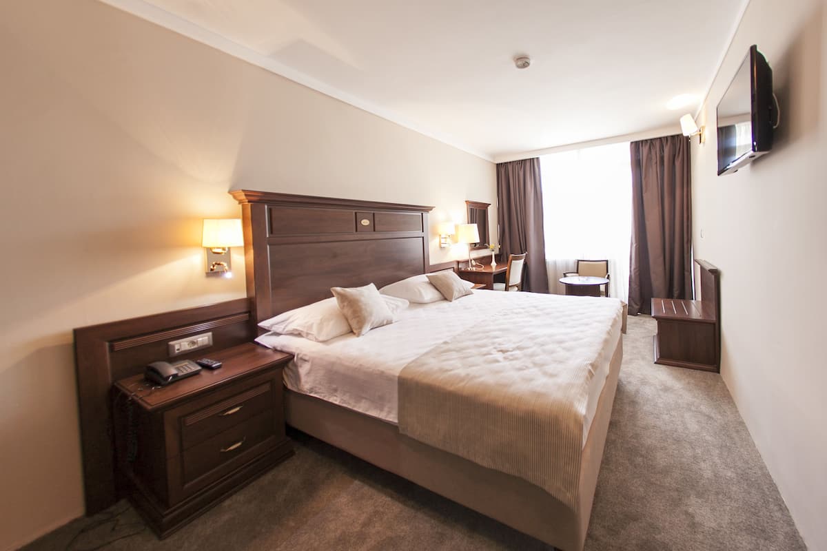 A hotel room with a large bed, bedside tables, a desk, a chair, a TV, and a window with curtains.