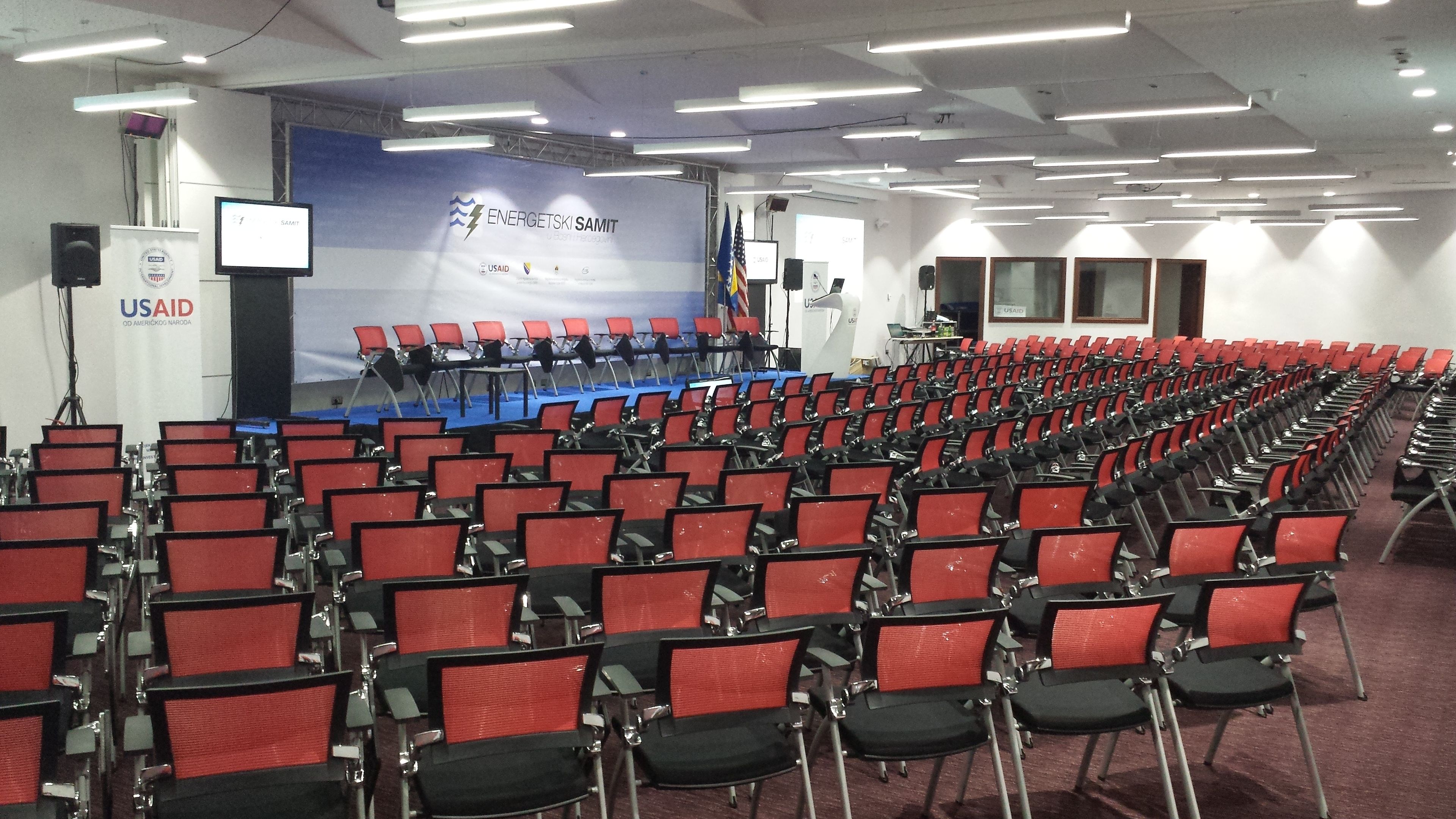 Ein Konferenzraum mit Reihen roter Stühle, einer Bühne mit blauem Teppich und USAID-Branding an den Wänden.