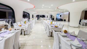 Een ruime balzaal met lange tafels gedekt voor een formeel evenement, met witte tafelkleden en stoelen, en een modern plafondontwerp.