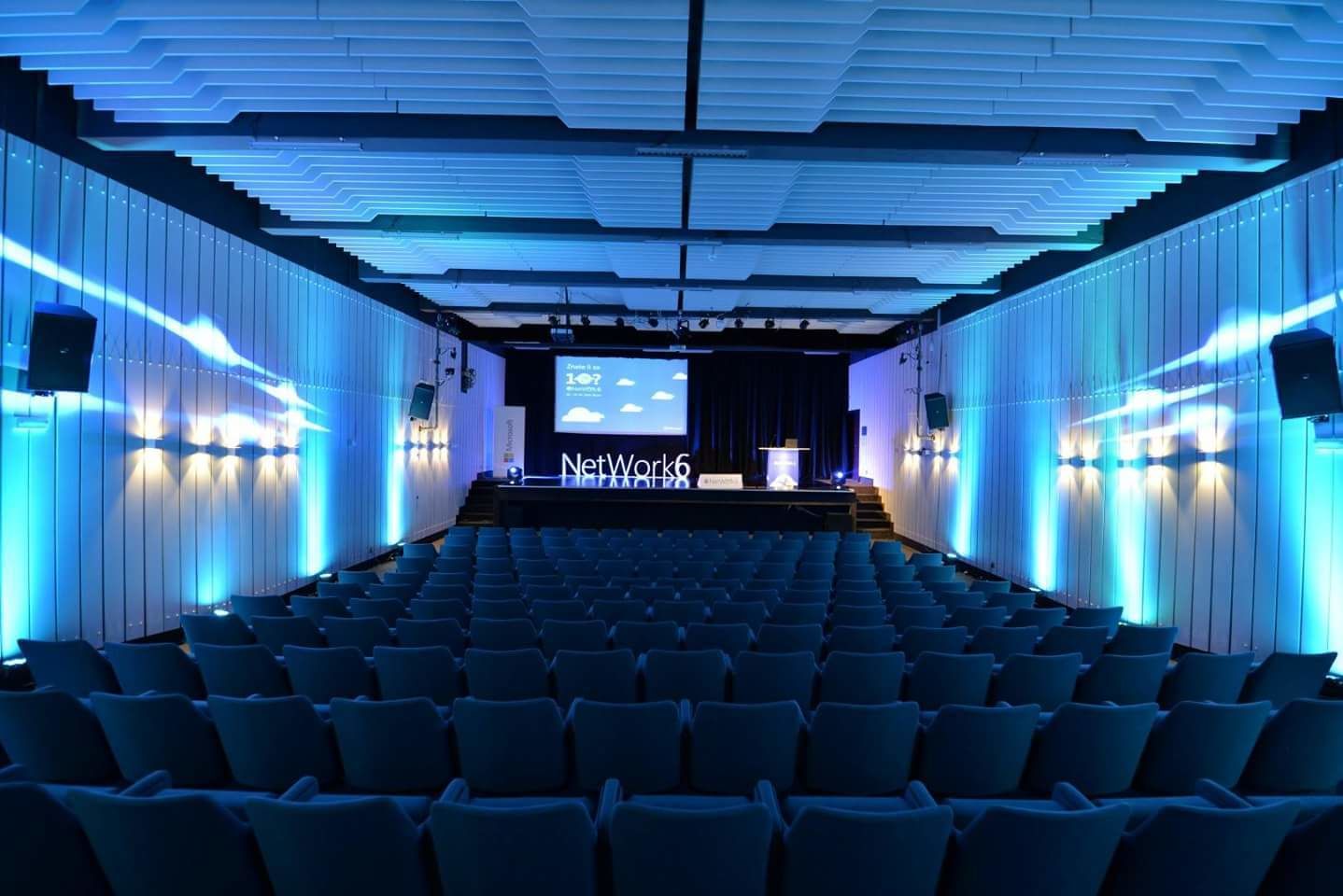 A conference room with rows of chairs, a stage with a presentation screen, and a logo for NetWork6.