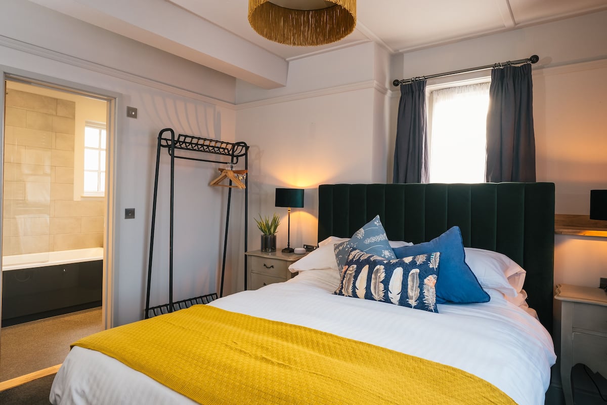 A bedroom with a bed, a bedside table, a nightstand, a window with curtains, a hanging light fixture, and a door leading to a bathroom.