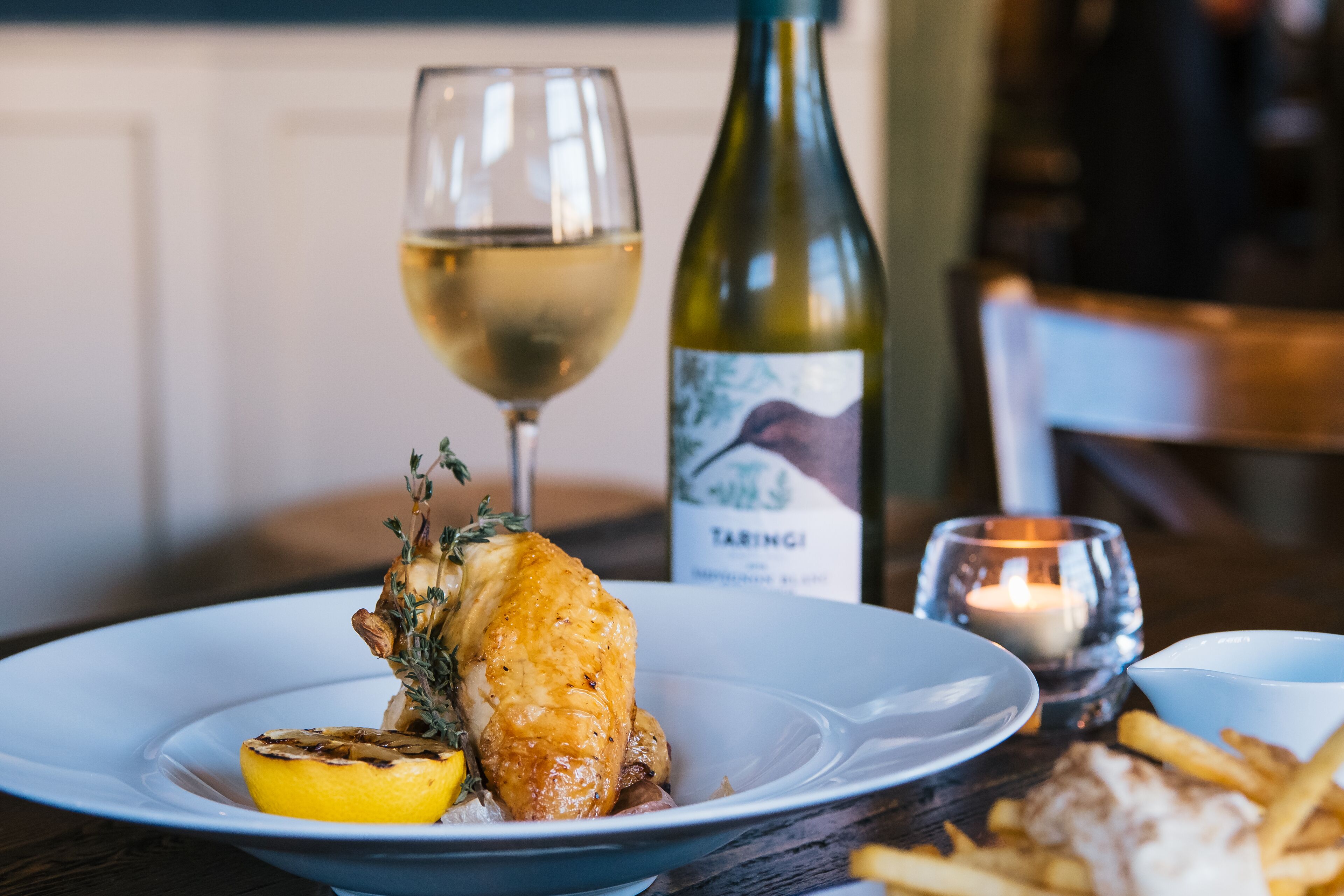 A plate with roasted chicken, lemon, and herbs, a glass of white wine, a bottle of wine, and a candle on a wooden table.