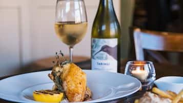 A plate with roasted chicken, lemon, and herbs, a glass of white wine, a bottle of wine, and a candle on a wooden table.