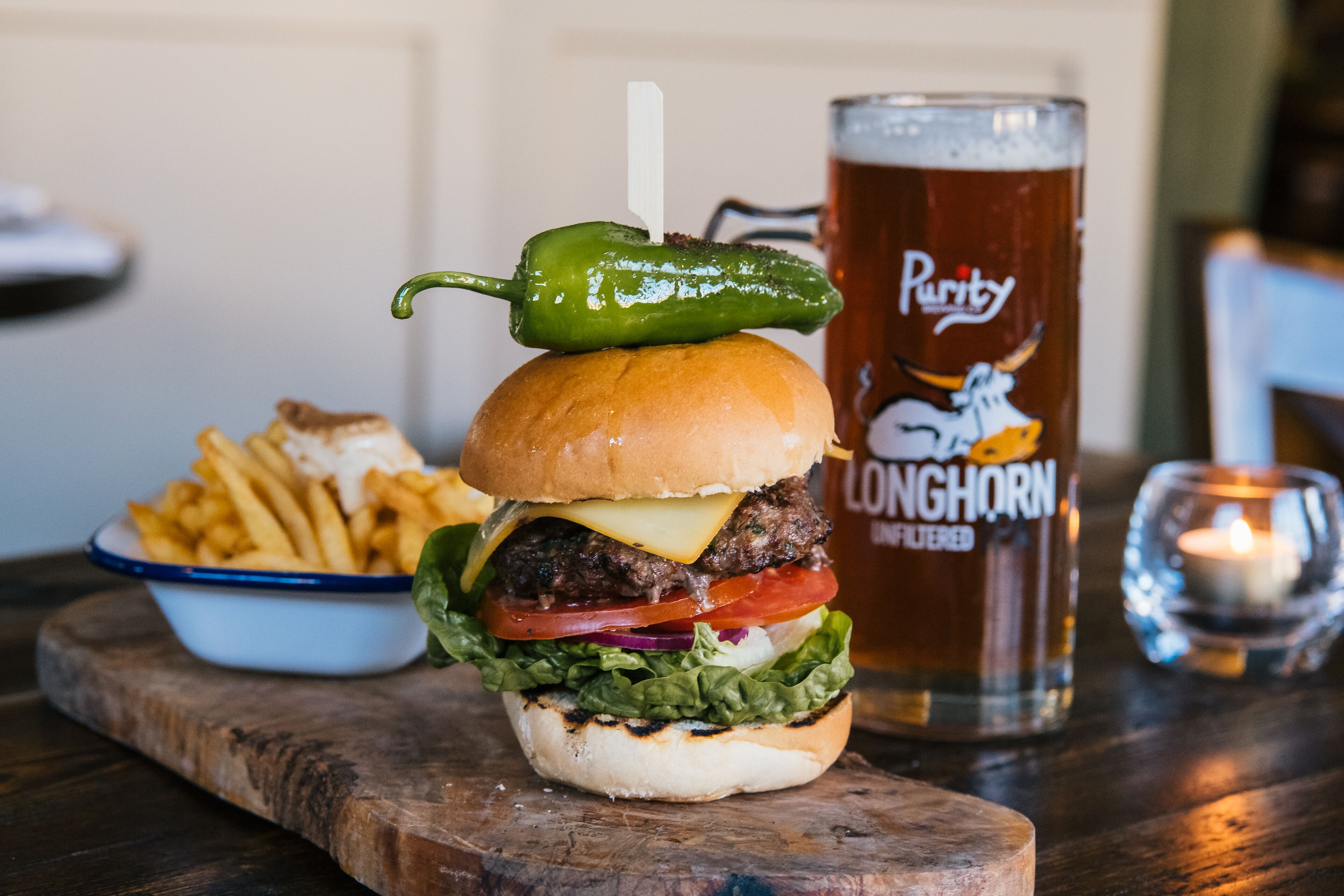 A burger with a green chili on top, a glass of beer, and a bowl of fries on a wooden board.