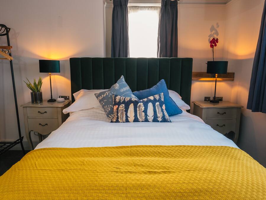 A neatly made bed with a yellow blanket, two matching pillows, and a dark headboard. There are two bedside tables with lamps and a small potted plant on one.