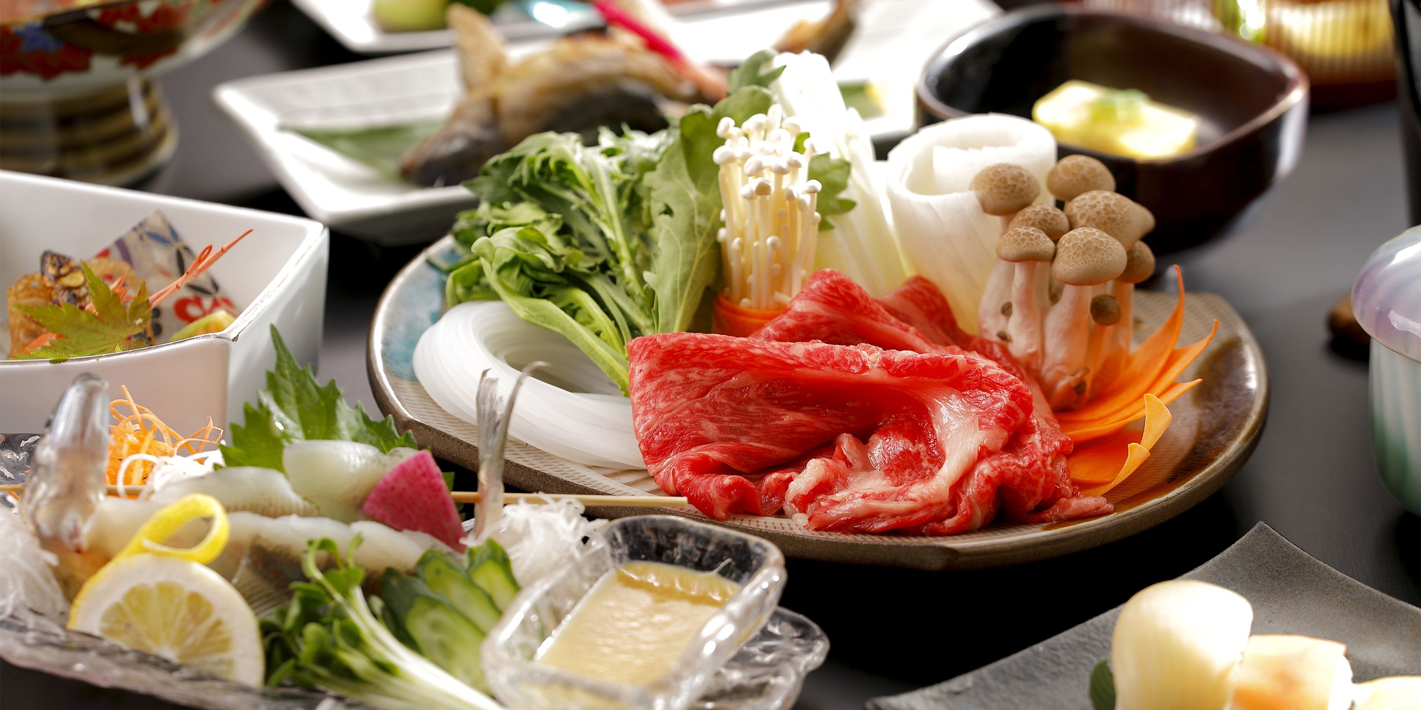 A variety of fresh ingredients and condiments on a table, including vegetables, mushrooms, and a dish with raw meat.