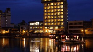 A multi-story hotel with illuminated windows, a dock with a boat, and a small building with a covered area. - Shokyoto No Yu Mikuma Hotel (Hita)