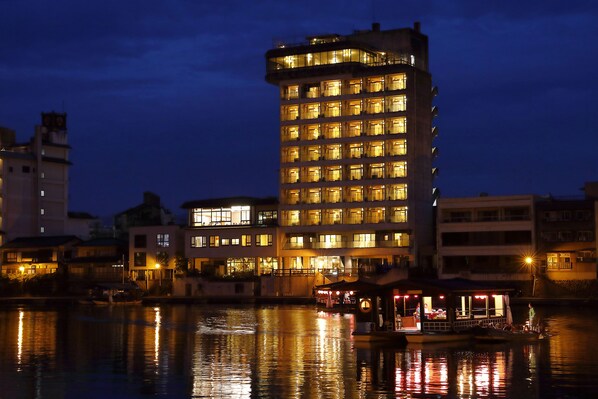 A multi-story hotel with illuminated windows, a dock with a boat, and a small building with a covered area. - Shokyoto No Yu Mikuma Hotel (Hita)