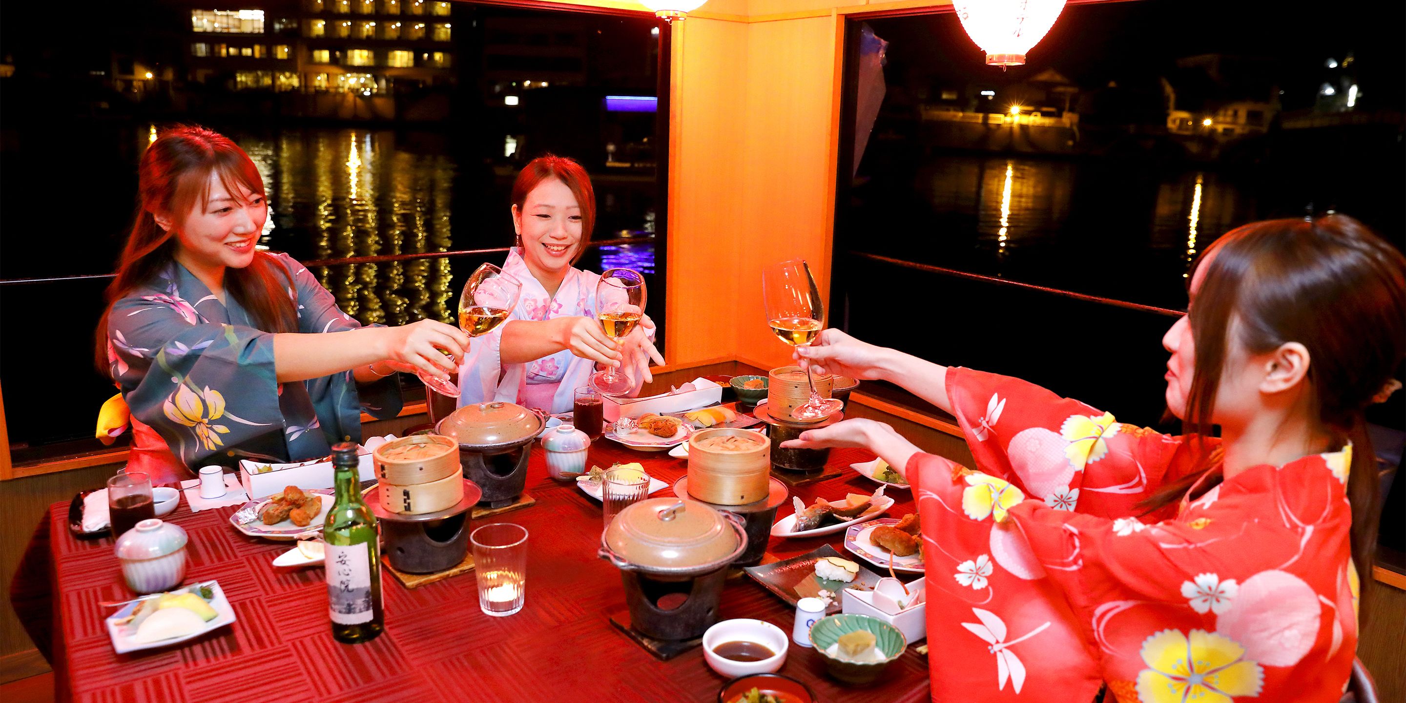 A group of people toasting with drinks at a table set with various dishes and a bottle of wine.