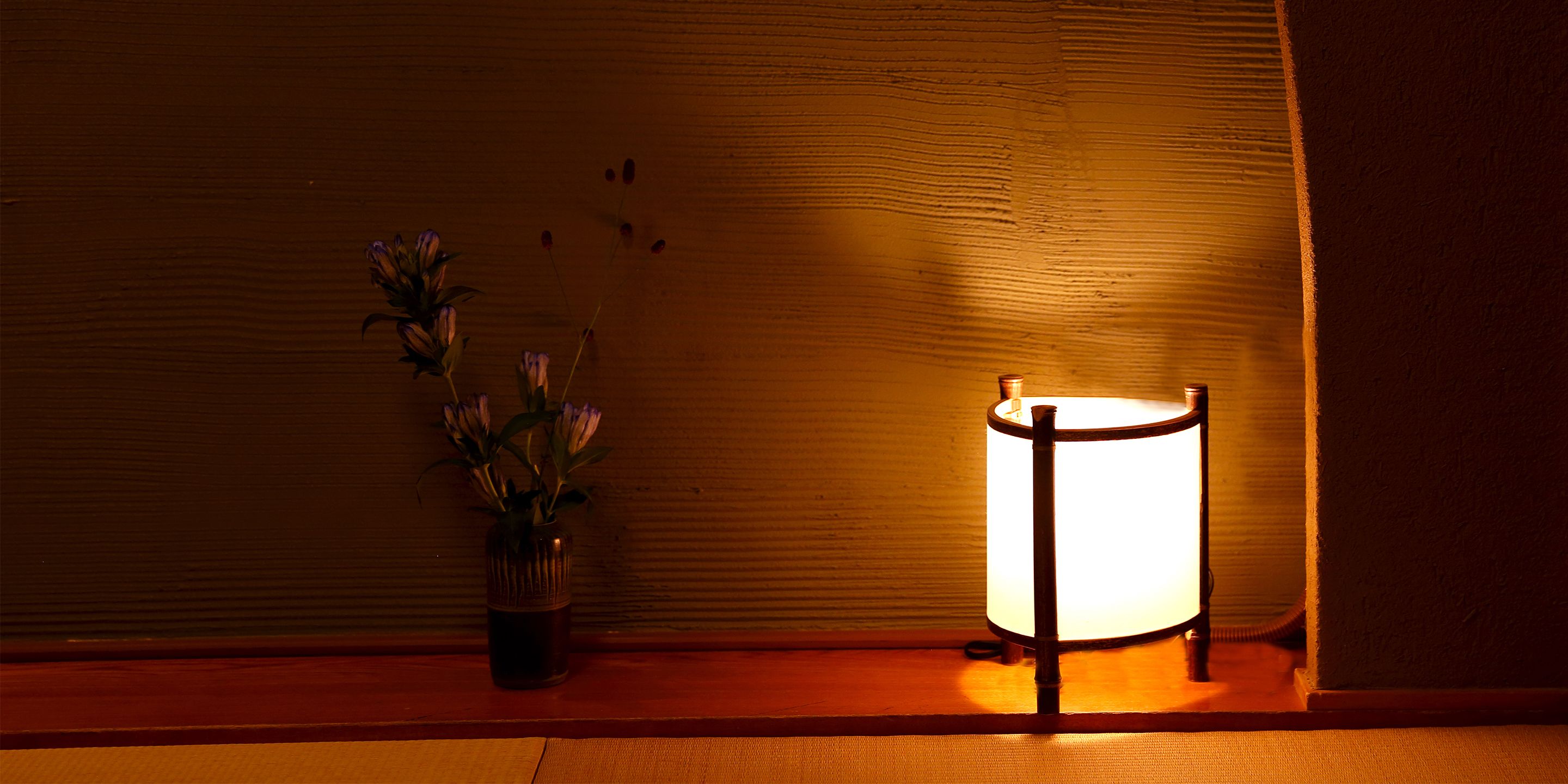 A lamp with a cylindrical shade on a wooden surface, a vase with flowers, and a textured wall in the background.