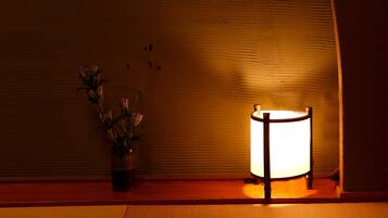 A lamp with a cylindrical shade on a wooden surface, a vase with flowers, and a textured wall in the background.