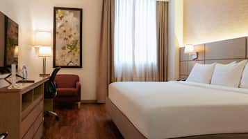 A hotel room with a bed, a desk with a lamp, a chair, and a large framed picture on the wall.