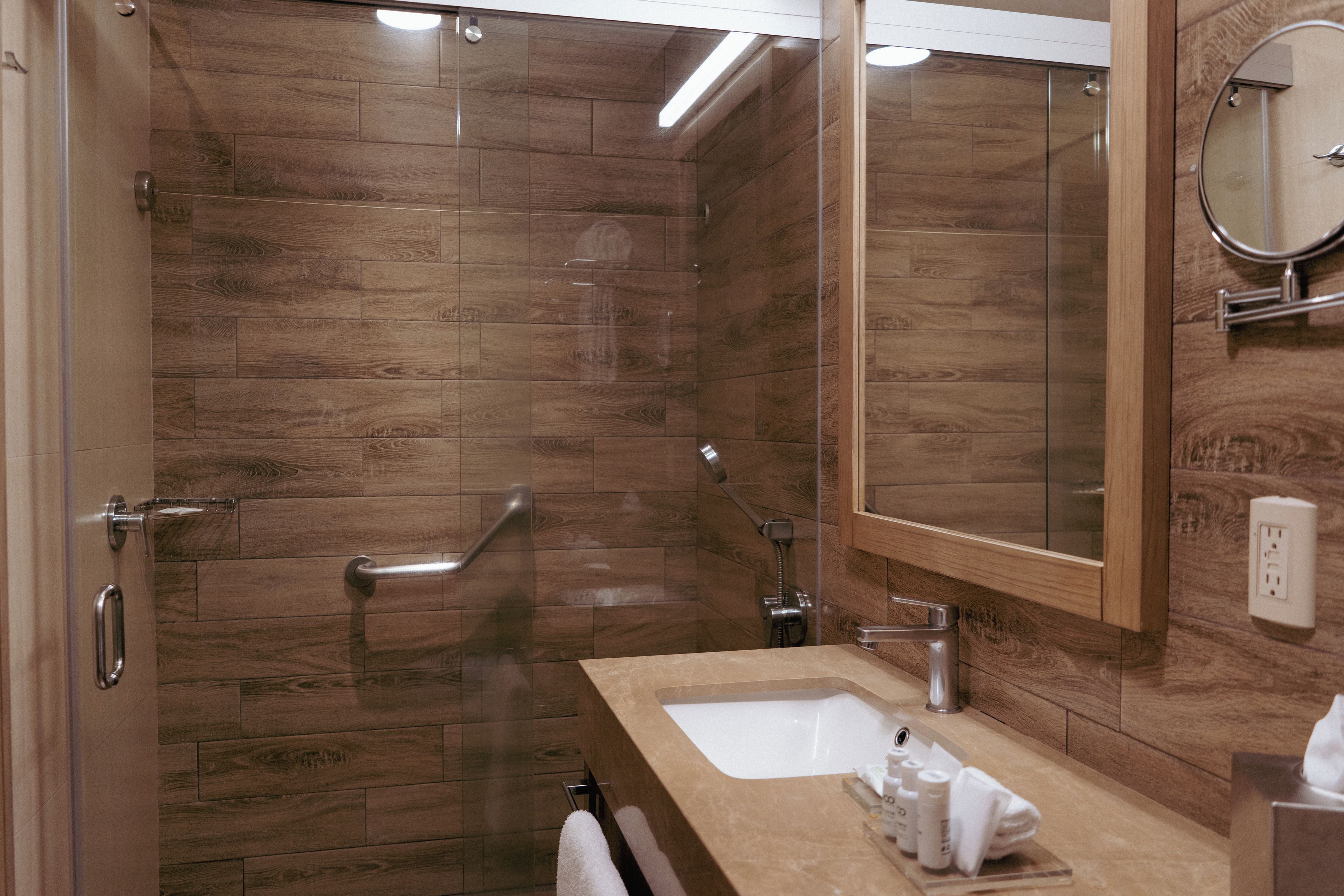 Ein Badezimmer mit Holzboden, einer Glasdusche, einem Waschbecken mit Spiegel und einem Handtuchhalter.