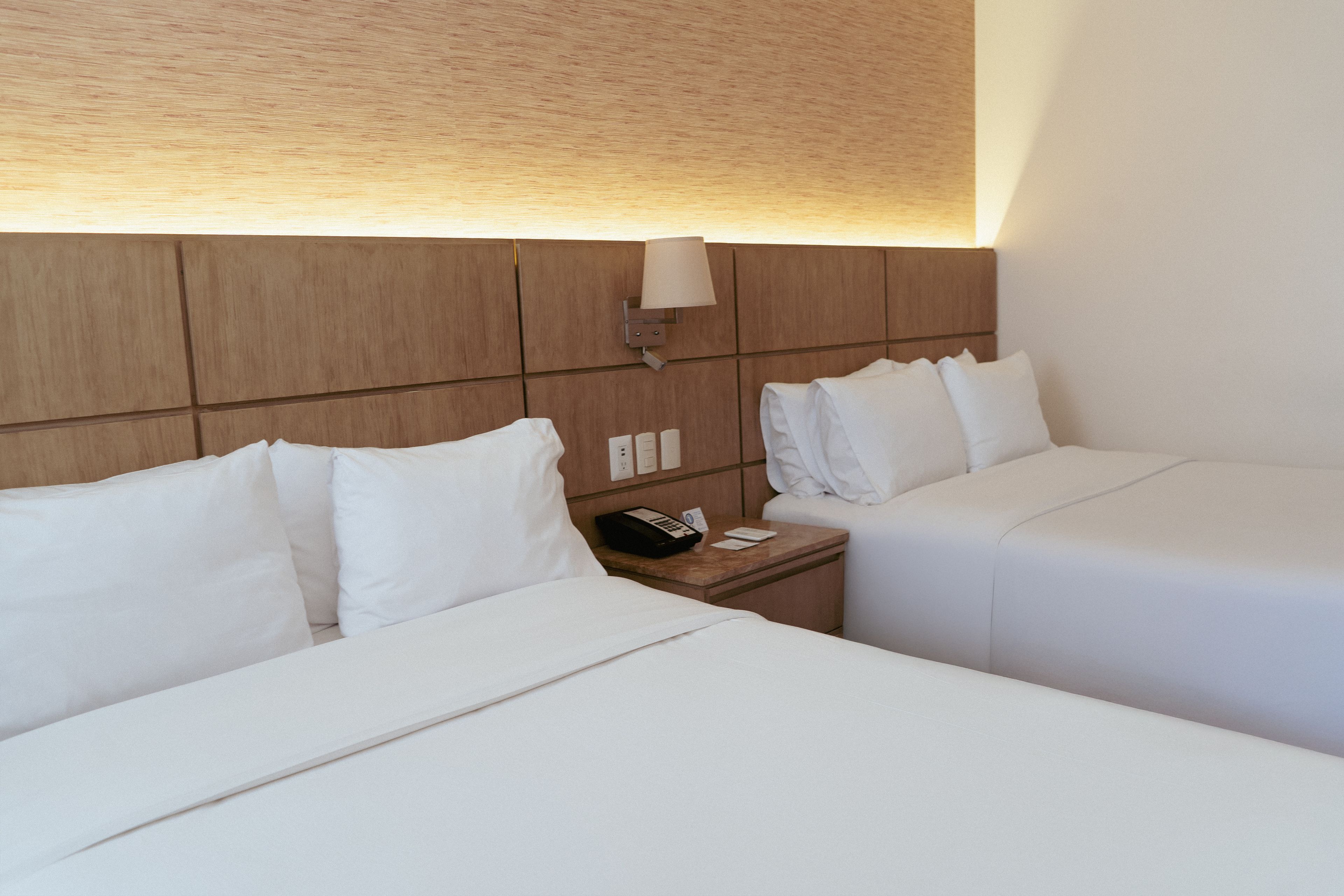 A hotel room with two single beds, a nightstand with a phone and a lamp, and a wooden headboard.