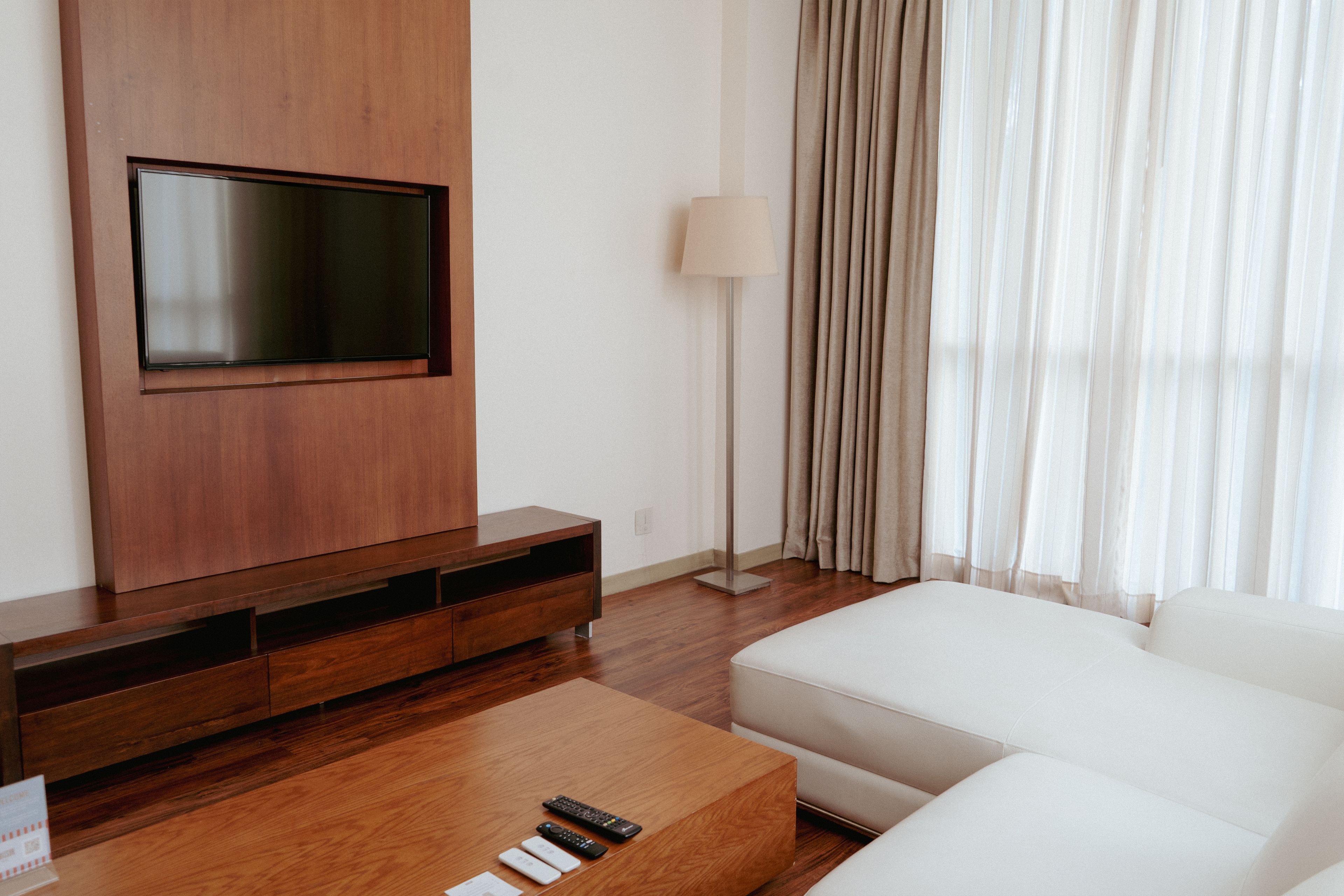 Una sala moderna con una televisión de pantalla plana montada en un mueble de madera, una lámpara de pie, un sofá blanco y una mesa de centro con controles remotos.