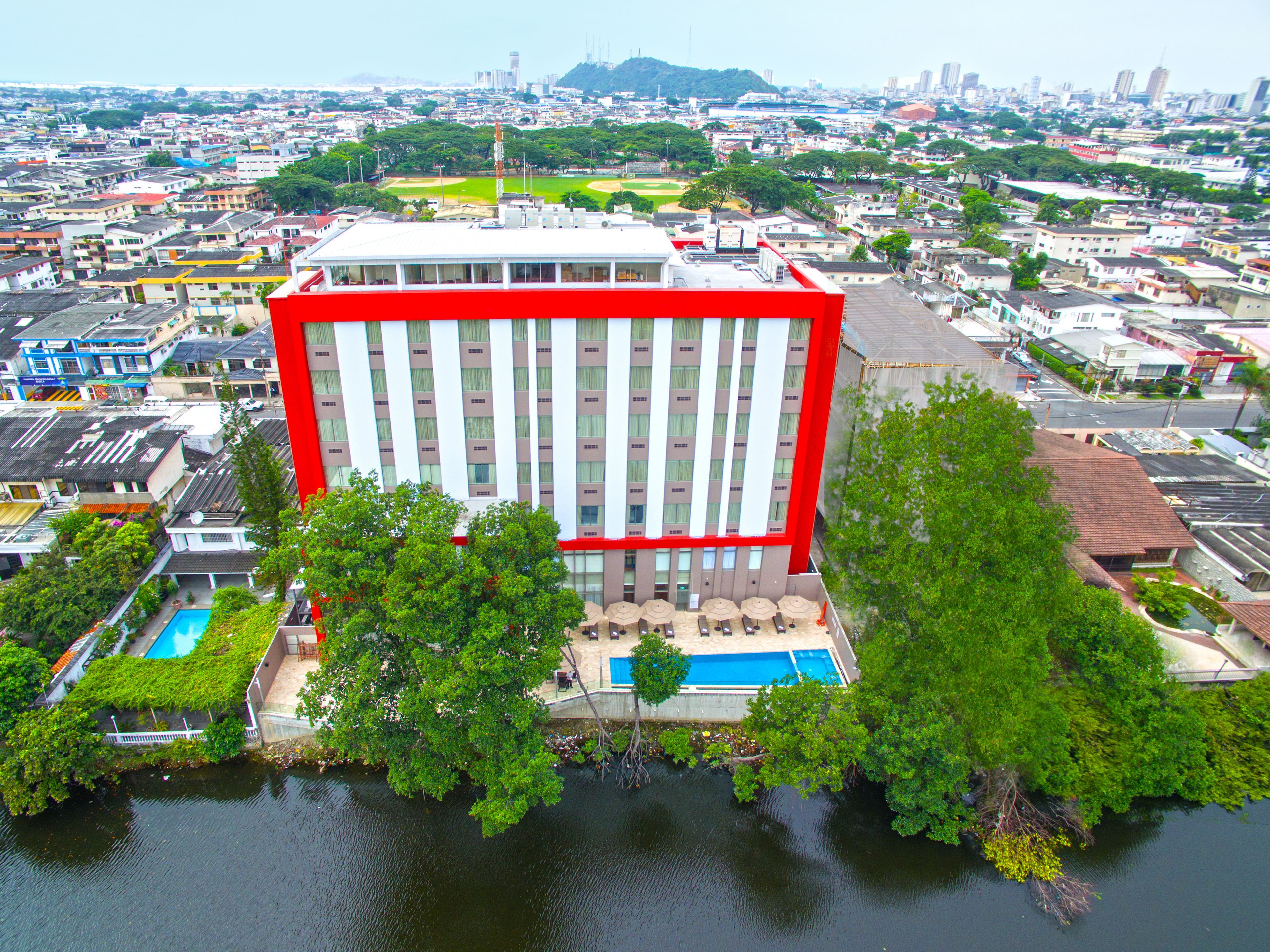 Een hoog hotel met een gevel in rood en wit, een zwembad op het dak en weelderige beplanting rondom het gebouw.