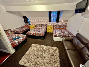 Un dormitorio compacto con tres camas, un sofá de cuero marrón, una alfombra pequeña y un televisor montado en la pared.