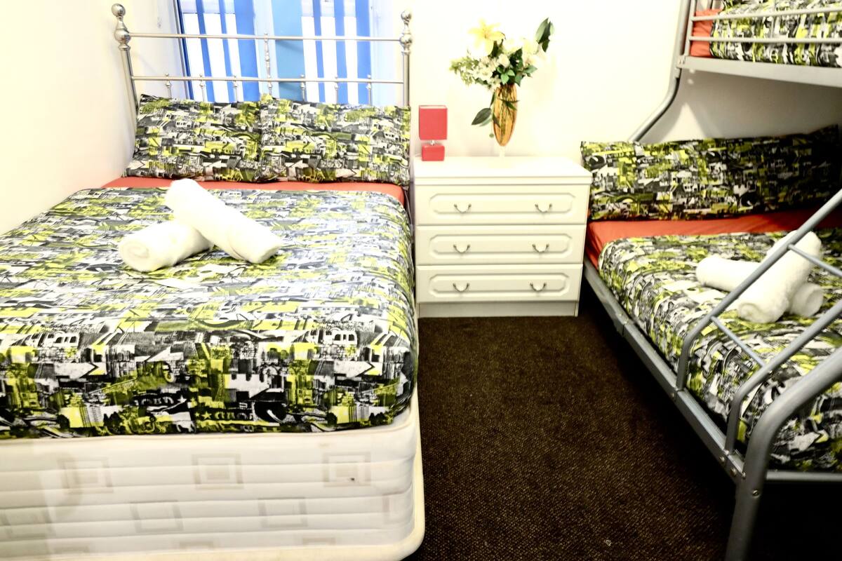 A dormitory room with two beds, each with patterned bedding and white towels. There is a small white dresser with drawers and a vase with flowers on top.