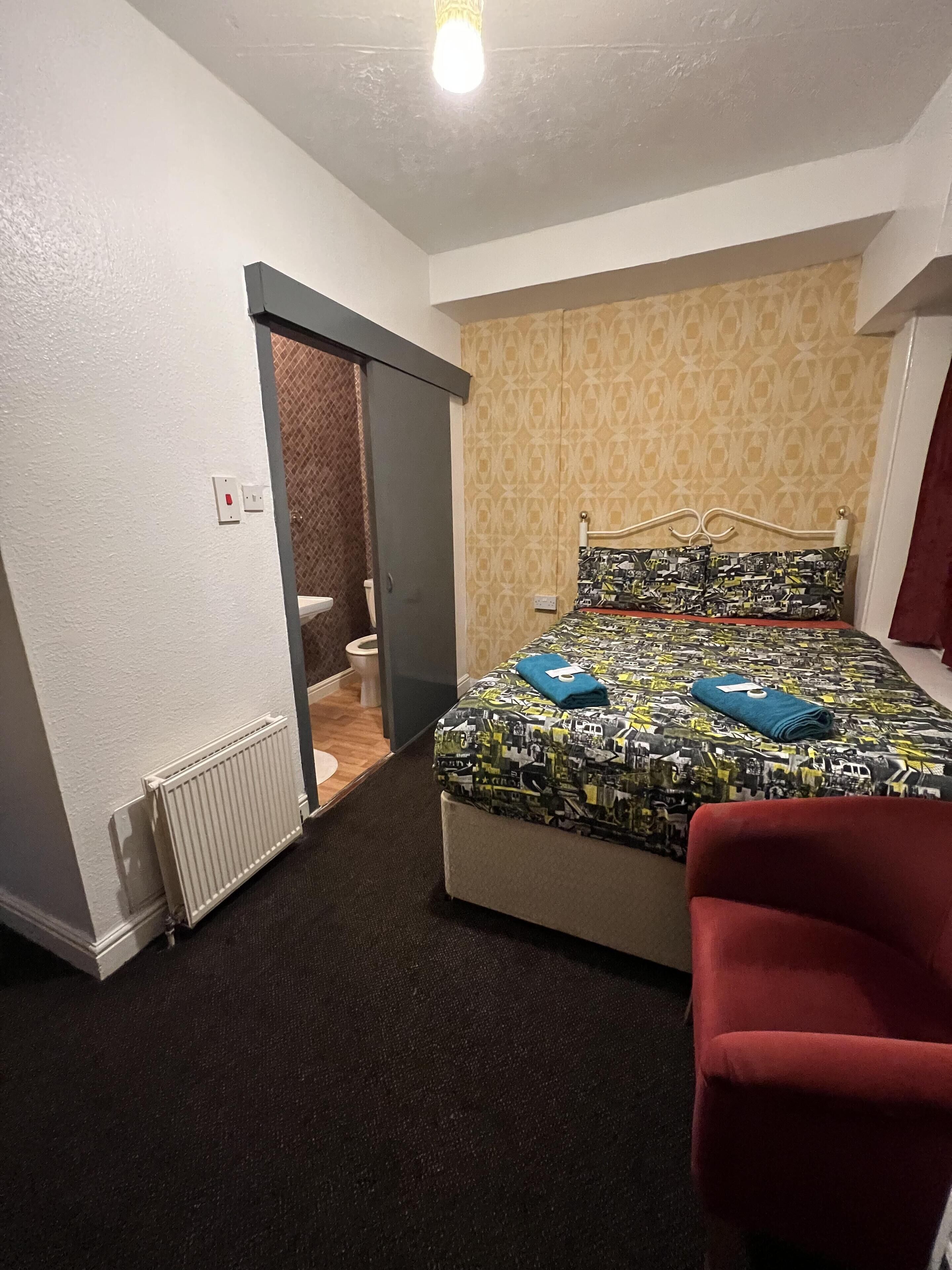 Ein Schlafzimmer mit einem Bett, einem roten Sessel, einem Heizkörper und einem durch eine offene Tür erreichbaren Badezimmer.