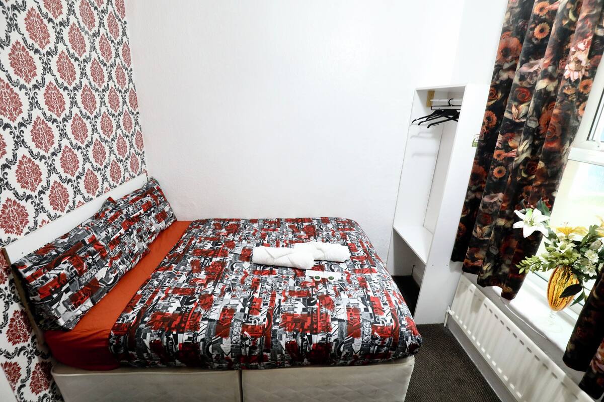 A small, neatly made bed with a patterned bedspread and a red pillow, a white radiator, and a window with floral curtains.