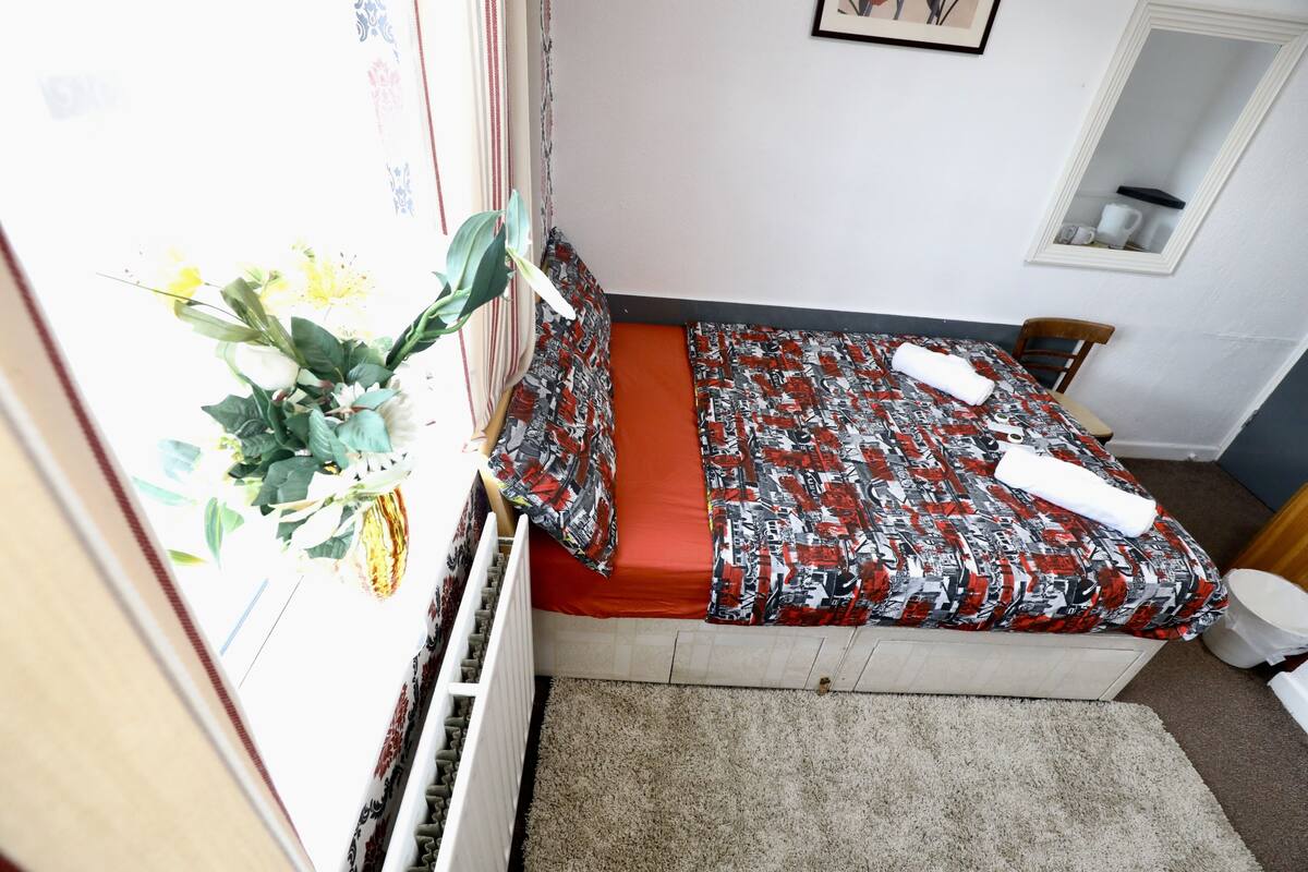 A small, single-bed room with a patterned bedspread, a window with curtains, a radiator, a small rug, and a chair.