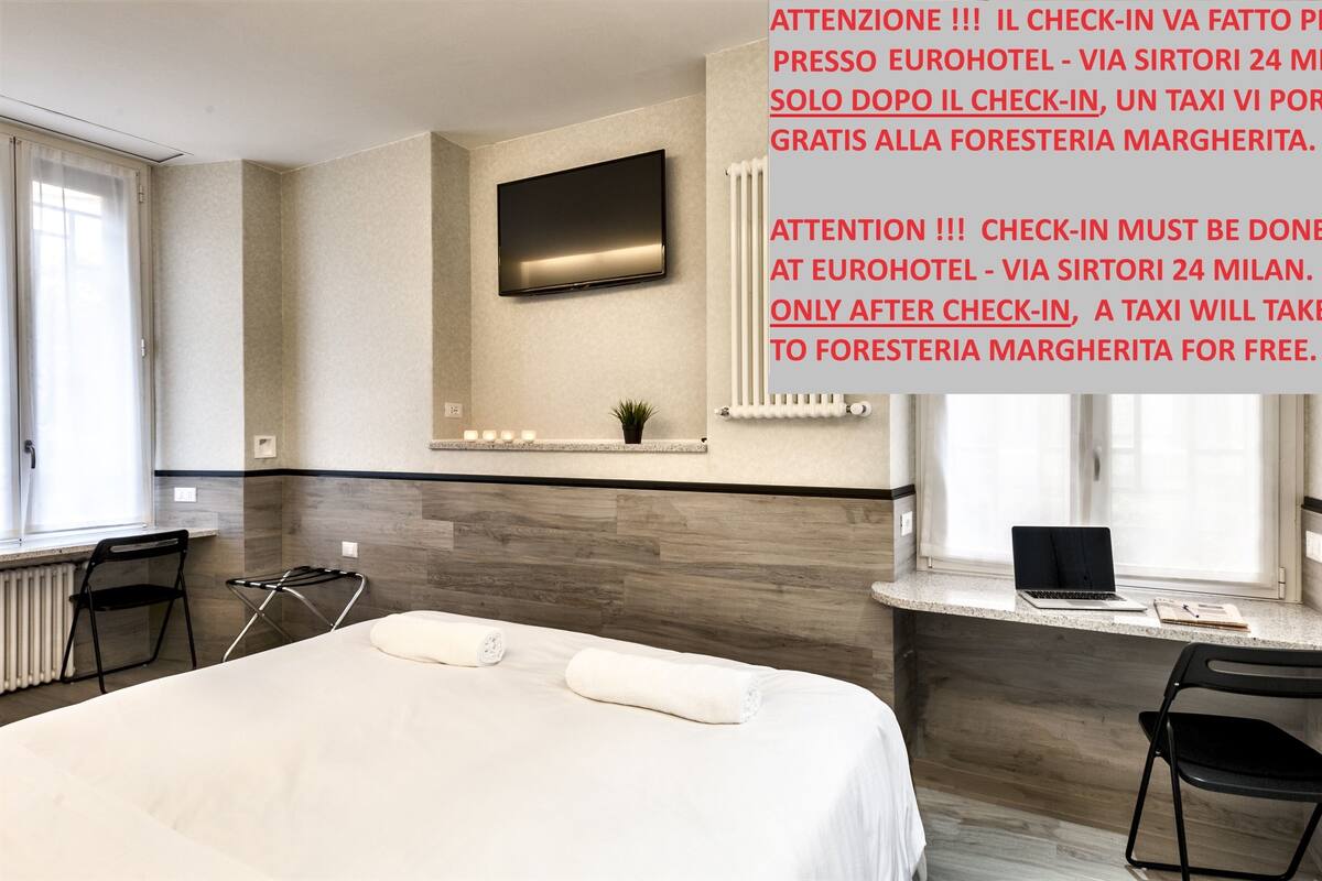A hotel room with a bed, a desk with a laptop, a chair, a TV mounted on the wall, and a window with curtains.