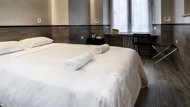 A modern hotel room with a large bed, a bedside table with a lamp, a window with curtains, and a radiator. - Foresteria Margherita Milano (Milan)